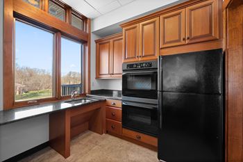 A kitchen with a black refrigerator, wooden cabinets, and a window overlooking a field.